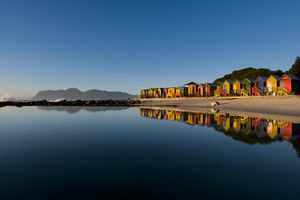 Row of colorful beach huts by a calm water body mountains in the background