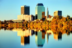 Cityscape with tall buildings reflected in a river trees lining the shore