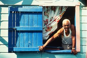 An elderly man in a white tank top leaning out of a colorful wooden house window with blue shutters