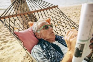 A person relaxing on a hammock at a beach while reading a newspaper