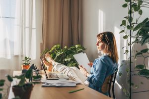 Woman sitting on chair with legs up on desk calculating bills with a laptop open and papers in hand.