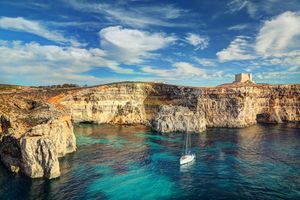A cliffside coastal landscape with a sailboat on the blue water and a tower in the background under a sunny sky
